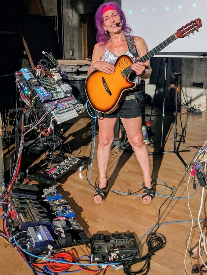 Rejyna holding a guitar beside a floor of looping pedals and cables